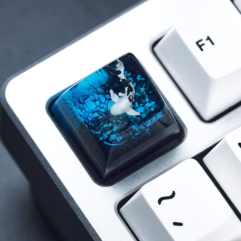 May include: A custom keyboard keycap featuring a koi fish design. The keycap has a clear resin top with a blue and white underwater scene. The koi fish is white with black markings. The keycap is square and sits among white keyboard keys.