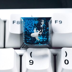 May include: A custom keyboard keycap featuring a koi fish design. The keycap is clear with a blue and white color scheme, with a koi fish swimming in a sea of blue. The keycap is surrounded by white keyboard keys.