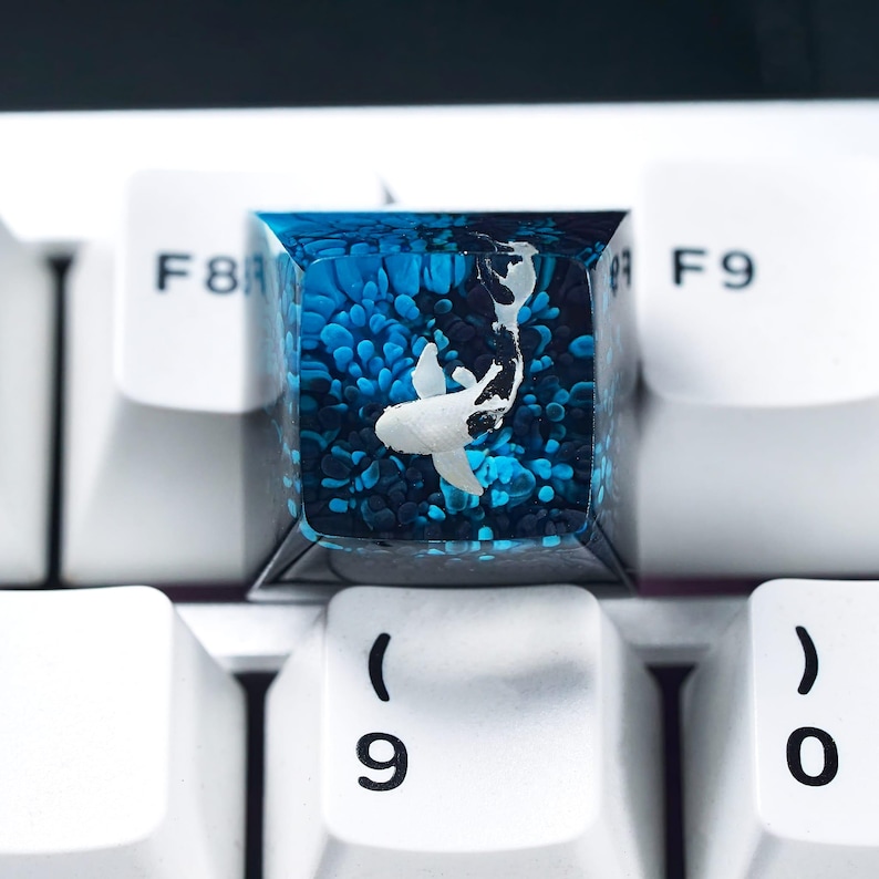 May include: A custom keyboard keycap featuring a koi fish design. The keycap is clear with a blue and white color scheme, with a koi fish swimming in a sea of blue. The keycap is surrounded by white keyboard keys.