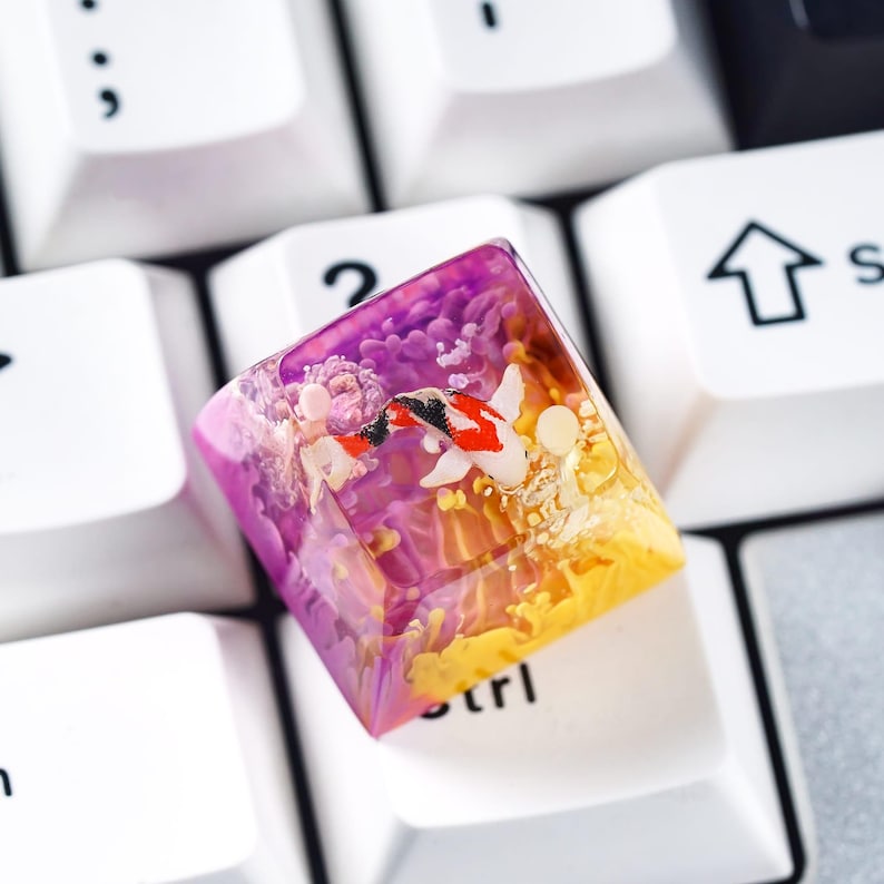 May include: A custom keycap featuring a koi fish design. The keycap is a square shape with a clear resin exterior. The interior design features a koi fish with red, black, and white markings, set against a purple and yellow background.