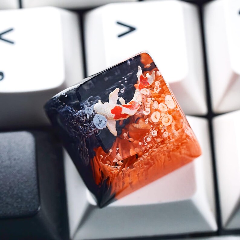 May include: A custom keycap featuring a koi fish design. The keycap has a clear resin body with a black and orange gradient, and a white, red, and black koi fish. The keycap is resting on a white keyboard.