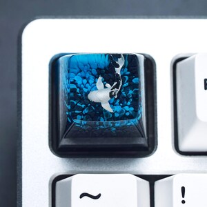 May include: A custom keyboard keycap featuring a koi fish design. The keycap is a square shape with a black base and a clear resin top. Inside the resin, a white and black koi fish swims in a blue and dark blue background.