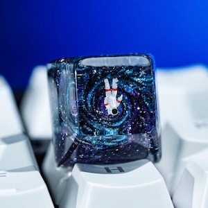 May include: A clear, square keycap with a miniature astronaut floating in a swirling galaxy design. The keycap is set against a background of white keyboard keys and a deep blue backdrop.