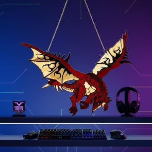 K&ouml;nnte beinhalten: Eine rote und schwarze Drachendekoration, die an einer Kette h&auml;ngt. Der Drache hat detaillierte Fl&uuml;gel und einen strukturierten K&ouml;rper. Ein kleines Schild mit dem Text "MONSTER HUNTER" befindet sich links. Gaming-Headset, Tastatur und Maus sind ebenfalls sichtbar.