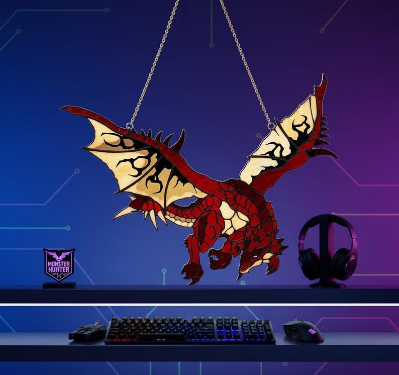 K&ouml;nnte beinhalten: Eine rote und schwarze Drachendekoration, die an einer Kette h&auml;ngt. Der Drache hat detaillierte Fl&uuml;gel und einen strukturierten K&ouml;rper. Ein kleines Schild mit dem Text "MONSTER HUNTER" befindet sich links. Gaming-Headset, Tastatur und Maus sind ebenfalls sichtbar.