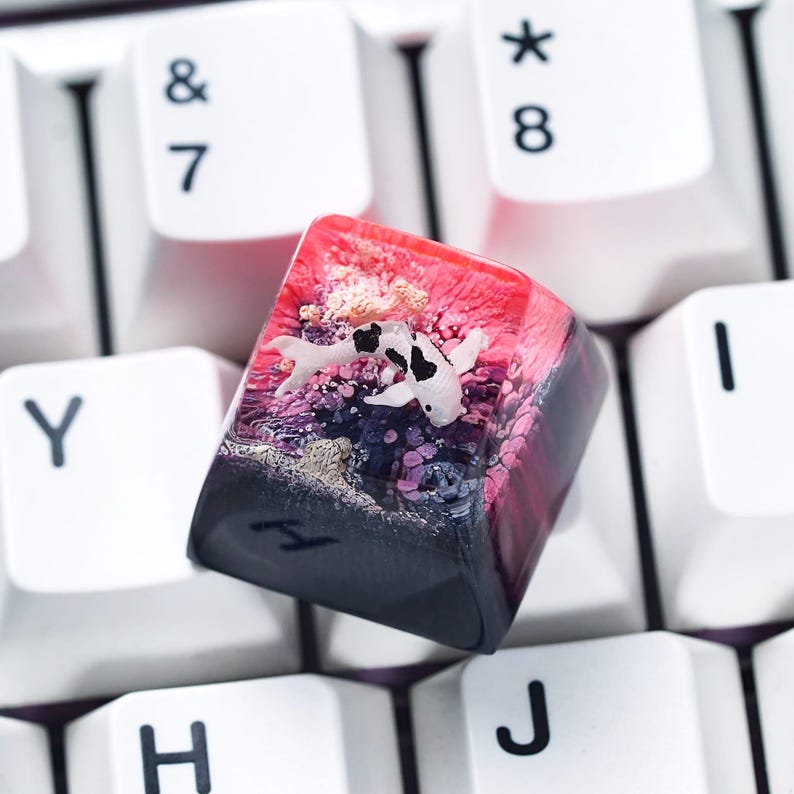 May include: A custom keyboard keycap featuring a koi fish design. The keycap is a mix of red, pink, black, and white. The koi fish is white with black spots, and the keycap has a glossy finish. The keycap is surrounded by white keyboard keys.