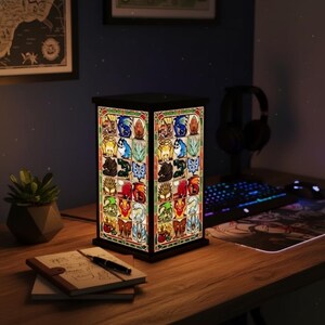 K&ouml;nnte beinhalten: Rechteckige Buntglaslampe mit schwarzem Rahmen. Die Lampe zeigt ein farbenfrohes, aufwendiges Design mit verschiedenen Fantasy-Motiven. Sie steht auf einem Holzschreibtisch neben einer kleinen Topfpflanze, einem Notizbuch und einem Stift.