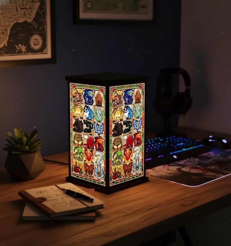 K&ouml;nnte beinhalten: Rechteckige Buntglaslampe mit schwarzem Rahmen. Die Lampe zeigt ein farbenfrohes, aufwendiges Design mit verschiedenen Fantasy-Motiven. Sie steht auf einem Holzschreibtisch neben einer kleinen Topfpflanze, einem Notizbuch und einem Stift.