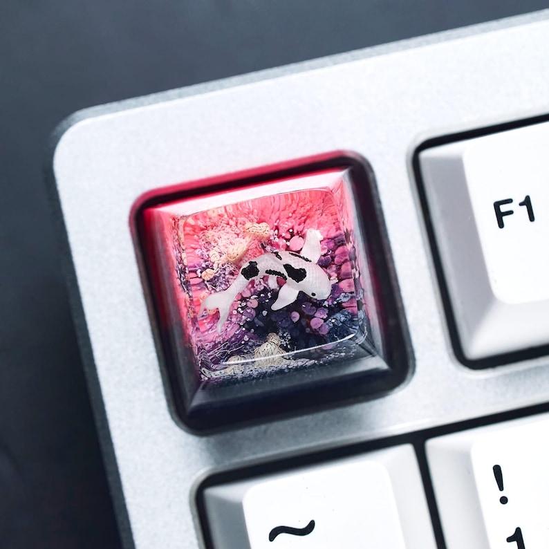 May include: A custom keyboard keycap featuring a koi fish design. The keycap is a square shape with a clear resin top, showcasing a white and black koi fish swimming over a colorful, textured background of pink, purple, and blue. The keycap is set on a silver keyboard.