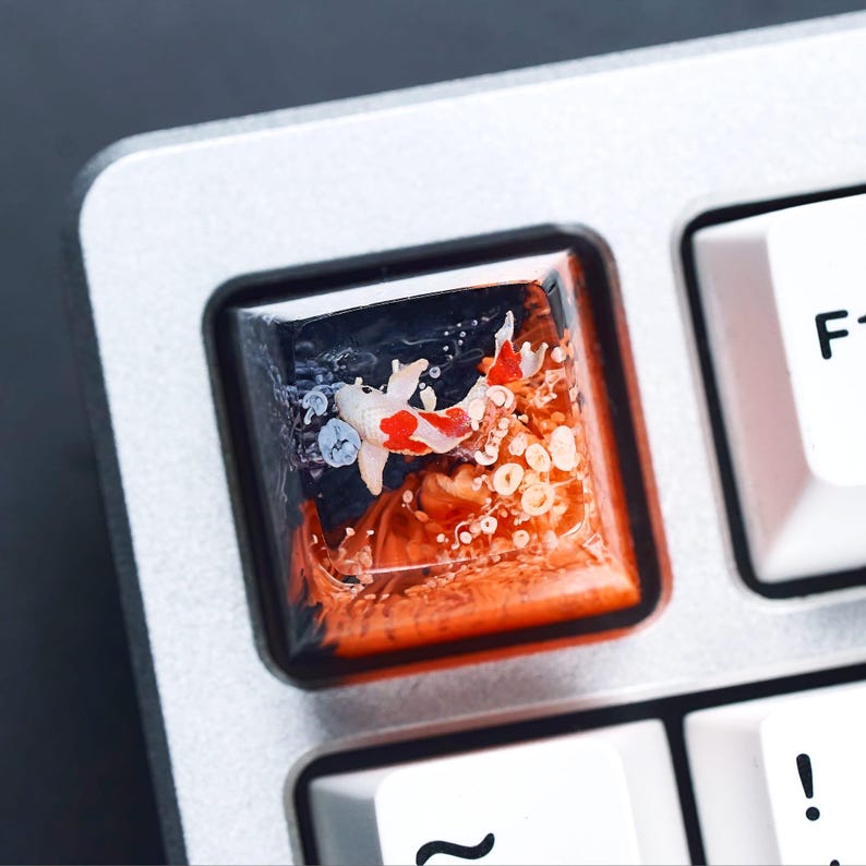 May include: A custom keyboard keycap featuring a koi fish design. The keycap is square and has a clear resin top with a detailed depiction of a koi fish swimming in a vibrant orange and blue environment. The keycap is set on a silver keyboard.