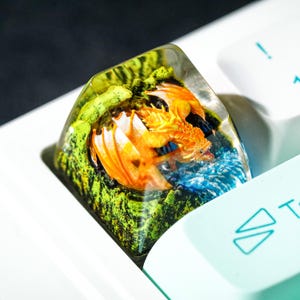 May include: A custom keycap featuring a detailed orange dragon set within a clear resin. The dragon is surrounded by green and blue elements, creating a miniature fantasy scene. The keycap is set on a white keyboard.