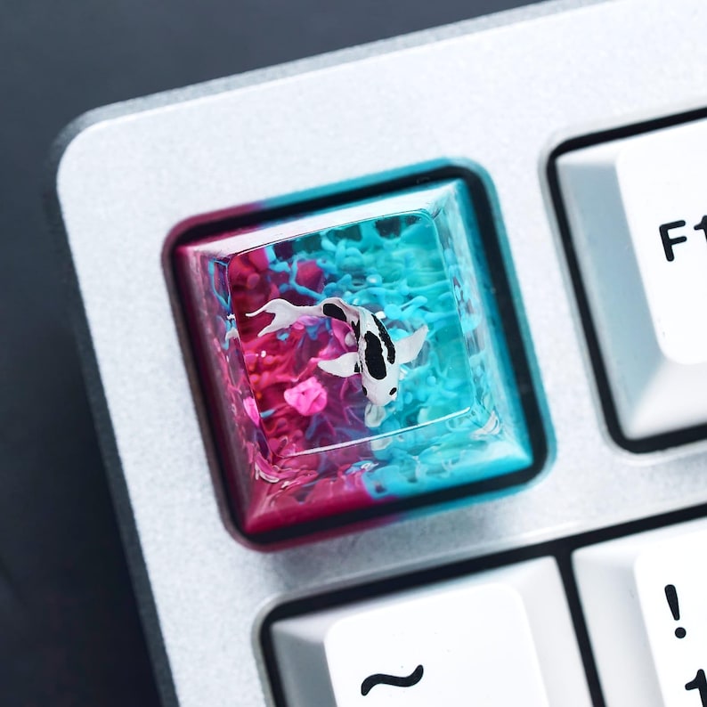 May include: A custom keyboard keycap featuring a koi fish design. The keycap is square, with a clear resin top that encases a black and white koi fish swimming in a vibrant, colorful background of pink and blue. The keycap is set on a silver keyboard.