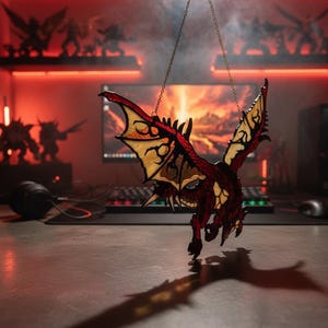 K&ouml;nnte beinhalten: Ein Drachenornament aus Buntglas, an einer Kette aufgeh&auml;ngt. Der Drache hat rote, gelbe und braune Farbt&ouml;ne mit detaillierten Fl&uuml;gelmustern. Das Ornament dient wahrscheinlich der Dekoration, m&ouml;glicherweise f&uuml;r einen Fantasy- oder Gaming-Enthusiasten.