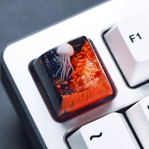 May include: A custom keyboard keycap featuring a jellyfish design. The keycap has a clear resin body with a white jellyfish and orange and dark blue accents. The keycap is surrounded by a silver keyboard frame and white keys.