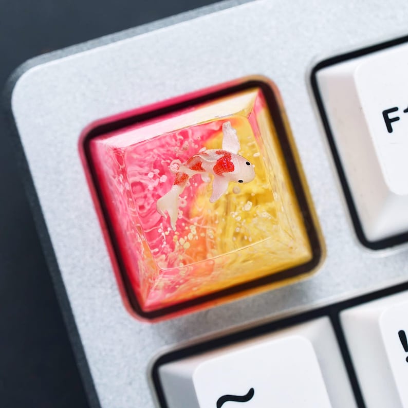 May include: A custom keyboard keycap featuring a miniature koi fish swimming in a clear resin enclosure. The keycap has a gradient of pink and yellow colors, with a red border. The keycap is set on a silver keyboard.