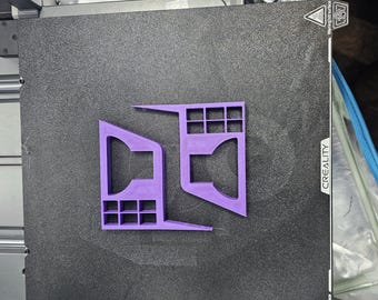 Puede incluir: Un objeto morado impreso en 3D con un diseño geométrico, con elementos rectangulares y angulados. El objeto está colocado sobre una superficie negra texturizada, posiblemente una base de impresora 3D. La palabra "CREALITY" es visible.