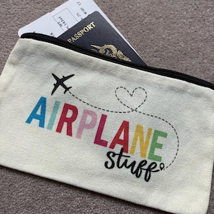 May include: Cream-colored travel pouch with the words "AIRPLANE stuff" in colorful letters and a black airplane graphic. A passport and boarding pass are visible inside the zippered pouch.