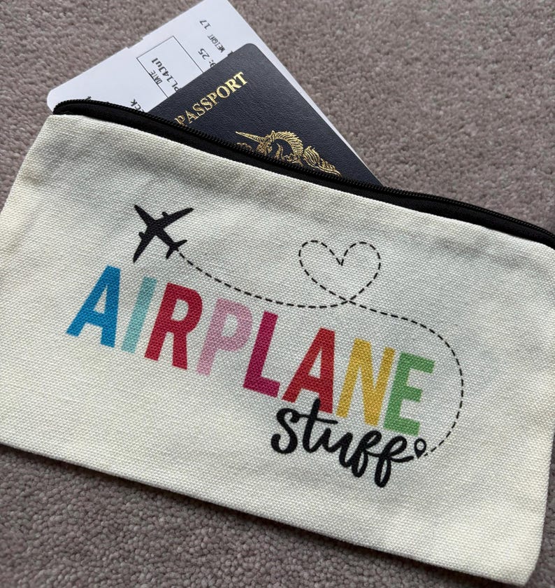 May include: Cream-colored travel pouch with the words "AIRPLANE stuff" in colorful letters and a black airplane graphic. A passport and boarding pass are visible inside the zippered pouch.