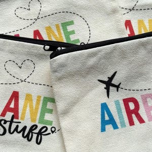 May include: Off-white canvas zipper pouches with black zippers. The pouches feature the words "AIRPLANE stuff" in colorful letters, with a dotted line and heart design. A black airplane graphic is also included.