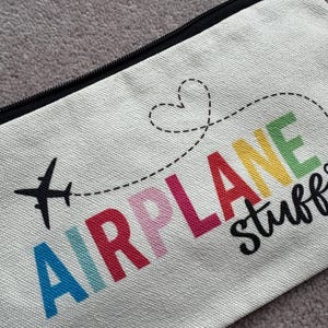 May include: Cream-colored zippered pouch with the words "AIRPLANE stuff" in colorful letters. A black airplane graphic and a dotted line with a heart shape are above the text. The pouch has a black zipper.