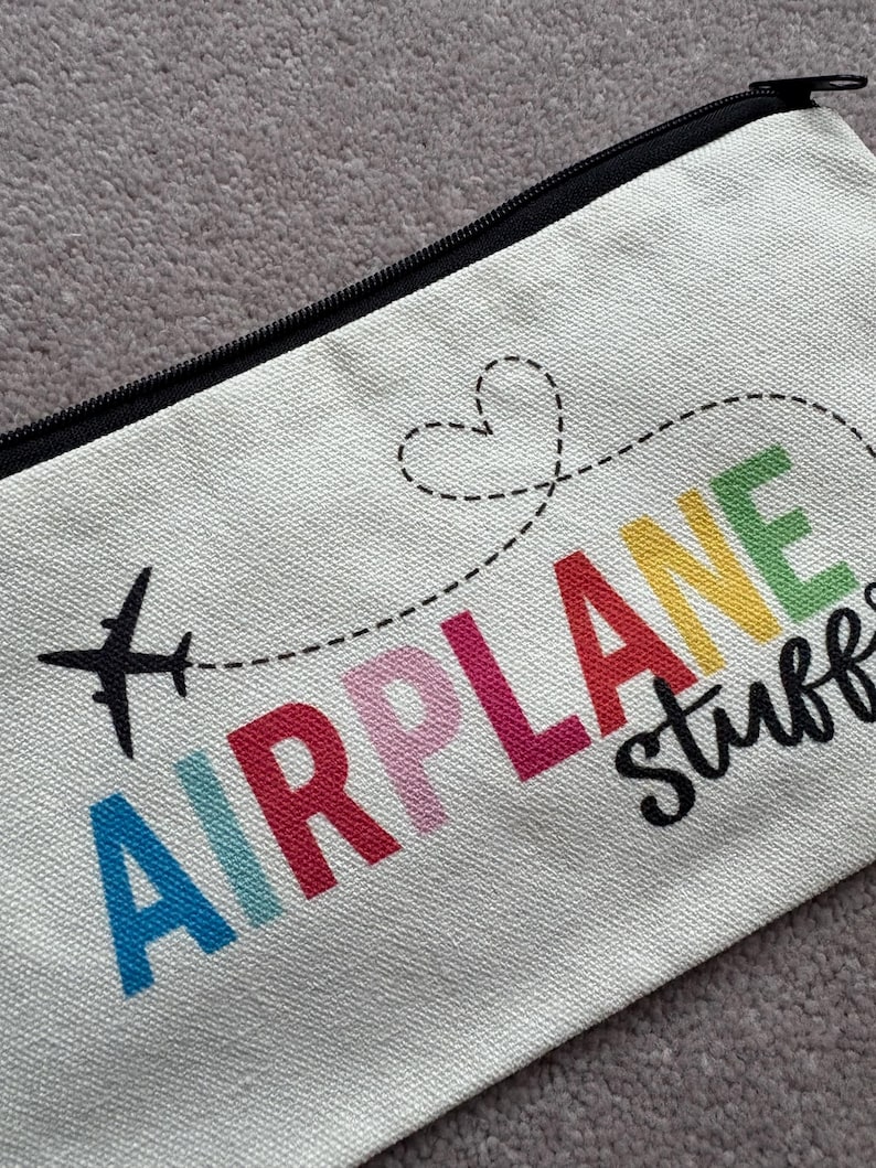 May include: Cream-colored zippered pouch with the words "AIRPLANE stuff" in colorful letters. A black airplane graphic and a dotted line with a heart shape are above the text. The pouch has a black zipper.