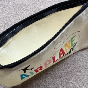 May include: Cream-colored pencil case with a black zipper and the words "AIRPLANE stuff" in colorful letters. A small black airplane graphic is also included. The interior is a light yellow color.