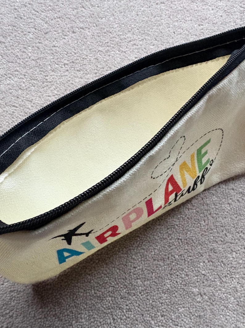May include: Cream-colored pencil case with a black zipper and the words "AIRPLANE stuff" in colorful letters. A small black airplane graphic is also included. The interior is a light yellow color.