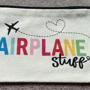 May include: A rectangular, off-white canvas pouch with a black zipper. The pouch features a black airplane graphic with a dotted line leading to the words "AIRPLANE stuff" in colorful letters.