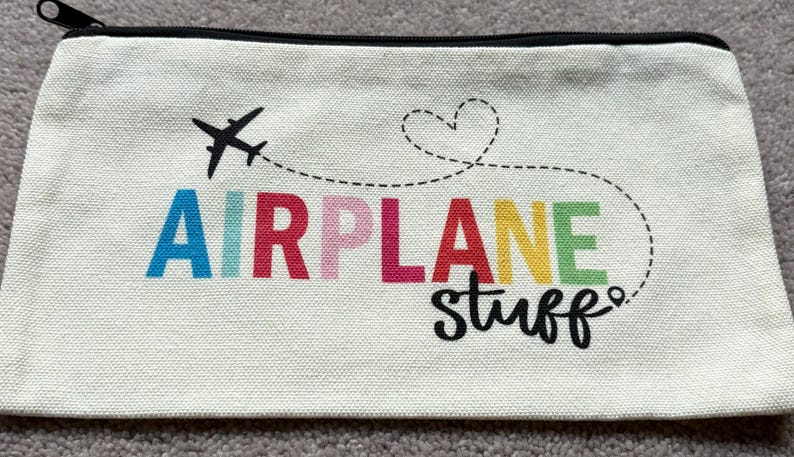 May include: A rectangular, off-white canvas pouch with a black zipper. The pouch features a black airplane graphic with a dotted line leading to the words "AIRPLANE stuff" in colorful letters.