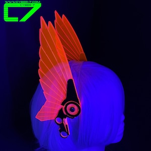 May include: A neon orange and black headpiece with wing-like structures, worn on a head with blue hair. The headpiece has a futuristic design with the text "WWW.CYBER-7.COM CYBER 7" in the upper left corner.