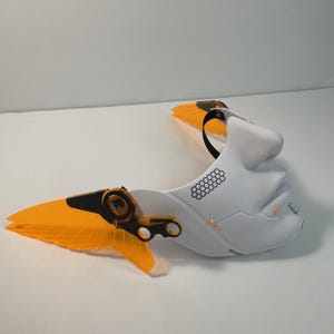 May include: White plastic mask with a human face design and orange wings. The mask has a black strap and a honeycomb pattern on the side.