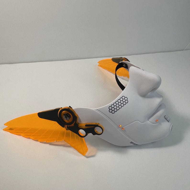 May include: White plastic mask with a human face design and orange wings. The mask has a black strap and a honeycomb pattern on the side.