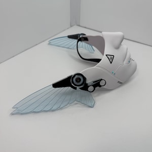 May include: A white and black futuristic headpiece with translucent blue wing-like structures. The headpiece has a curved design with a black strap and small blue accents. A triangular symbol is visible on the side.
