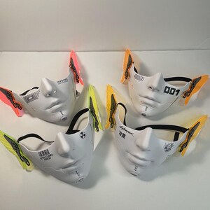 May include: Four white face masks with different designs and black straps. Each mask has a pair of colorful butterfly wings attached to the sides. The masks feature text such as "Cyberpunk", "001", and "Level 107 Area Access".