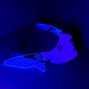 May include: A futuristic white and blue-lit headpiece with wing-like structures and a geometric emblem. The design features a sleek, modern aesthetic with glowing blue accents, suggesting a sci-fi or cyberpunk theme.