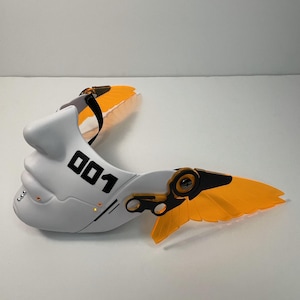 May include: White plastic mask with black details and the number "001" printed on the side. The mask has two orange wings attached to the sides.