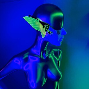 May include: A mannequin wearing a green and black futuristic-style face mask with a wing design. The mask has a barcode on the side. The mannequin is lit with blue and green light.