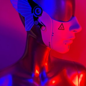May include: A futuristic headpiece with a white face mask and a blue wing-like structure. The mask has a hazard symbol and text. The mannequin is metallic and reflects red and blue light.