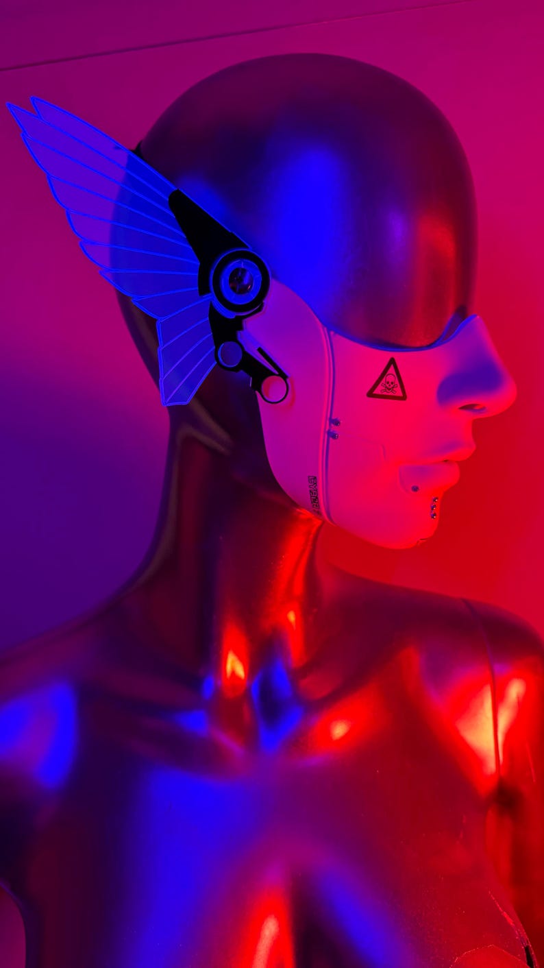 May include: A futuristic headpiece with a white face mask and a blue wing-like structure. The mask has a hazard symbol and text. The mannequin is metallic and reflects red and blue light.