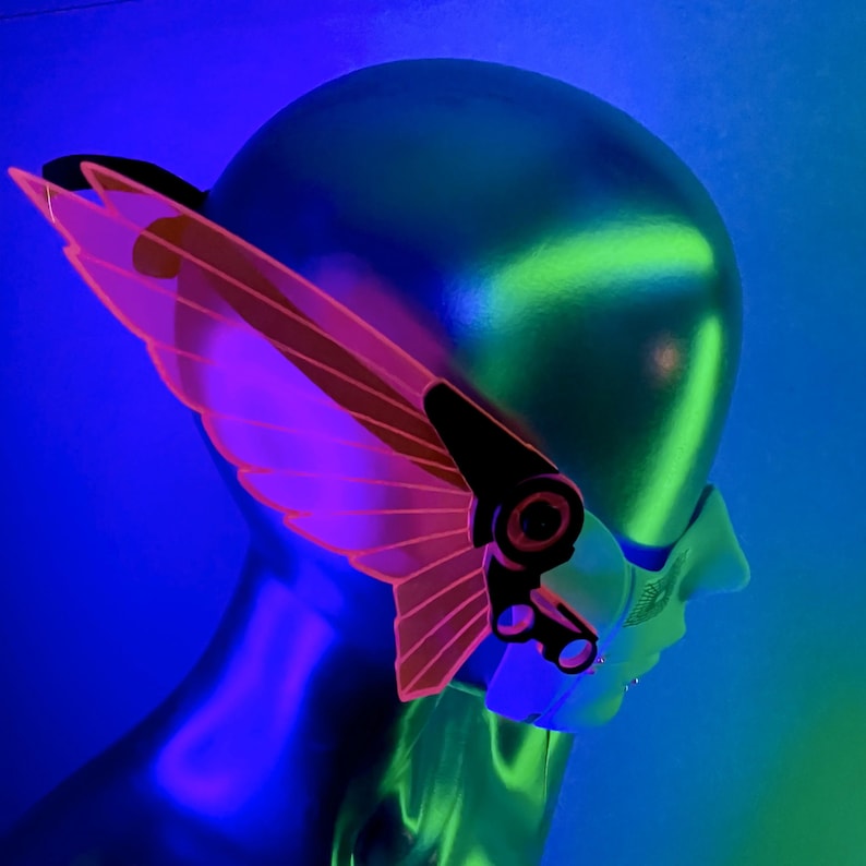 May include: A pink and black futuristic mask with a wing design. The mask is made of plastic and has a clear visor. The mask is being worn by a mannequin.