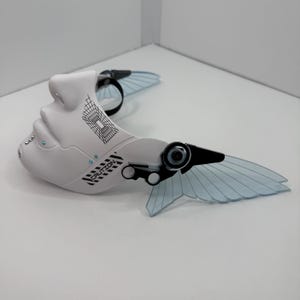 May include: A white, futuristic mask with a face-like design, featuring black and blue accents. The mask has a wing-like structure with transparent blue panels and the word "CAUTION" printed on it. The mask is designed with a black strap.