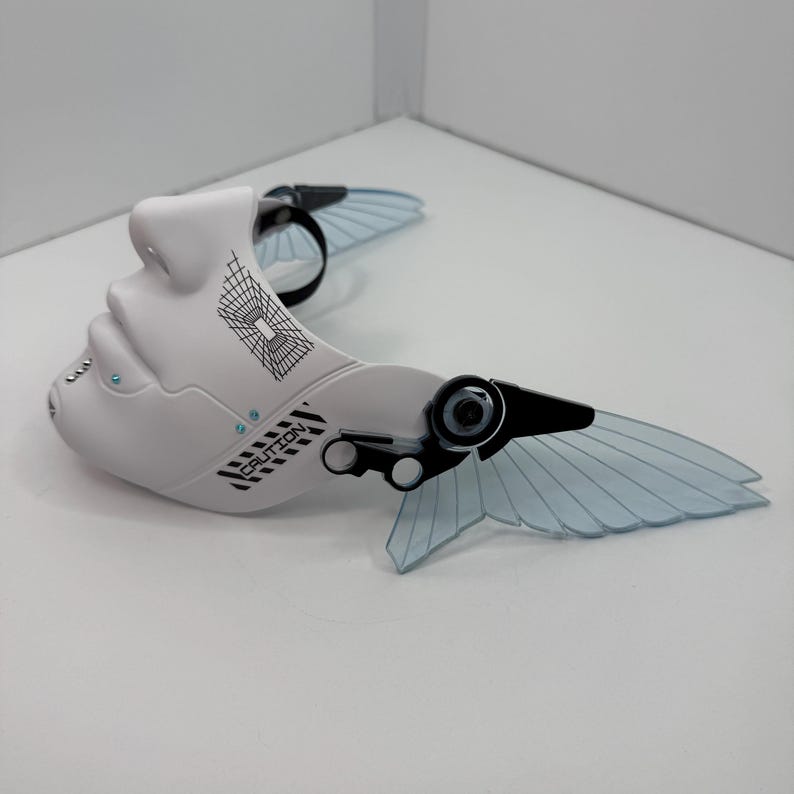 May include: A white, futuristic mask with a face-like design, featuring black and blue accents. The mask has a wing-like structure with transparent blue panels and the word "CAUTION" printed on it. The mask is designed with a black strap.