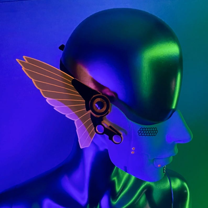 May include: A futuristic-looking mask with a glowing orange wing design. The mask is made of black plastic and has a silver metal accent. The mask is worn by a person with a blue and green background.