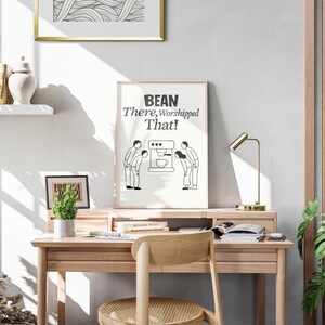 Peut inclure: Une affiche encadrée avec le texte "BEAN There, Worshipped That!" et une illustration de personnes s'inclinant devant une machine à café. L'affiche est exposée sur un bureau en bois avec une lampe de bureau dorée et une petite plante en pot.