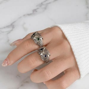 May include: Two silver-toned rings shaped like owl heads, each featuring textured feather detailing. One ring has small green gemstone eyes, while the other ring features dark blue gemstone eyes. The rings are worn on two fingers of a hand.