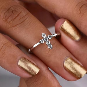 Peut inclure: Une bague en argent ornée d'une croix sertie de pierres précieuses claires et étincelantes. La croix est composée de cinq pierres rondes. La bague est portée sur un doigt vernis de couleur or.