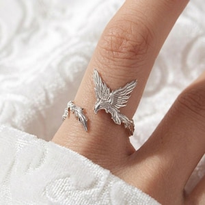 May include: A silver-colored ring featuring a detailed phoenix design. The ring wraps around the finger, with the phoenix's wings and head prominently displayed. The ring is worn on a finger against a soft, white background.