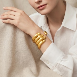 Può includere: Un bracciale rigido dorato a forma di mano con le dita che si avvolgono attorno al polso. Il bracciale presenta una finitura liscia e opaca e un design scultoreo che imita la forma delle dita, creando una fascia continua attorno al braccio.