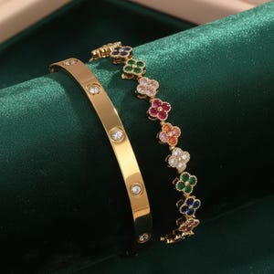 May include: Two gold bracelets. One is a solid band with clear, round gemstones. The other is a chain of colourful, four-leaf clover charms. The charms are in shades of pink, orange, green, blue, and white.