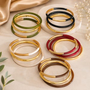 Puede incluir: Una colección de brazaletes circulares dispuestos en cinco grupos distintos. Cada brazalete presenta un diseño bicolor, combinando un segmento de metal pulido de color dorado con un segmento de resina lisa de color sólido en verde oliva, negro, blanco, rojo o marrón.
